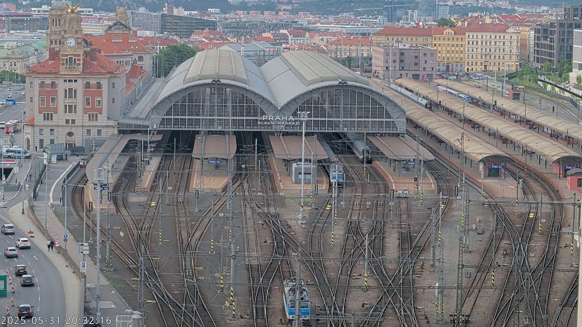 An aerial view of a large train station with numerous tracks and platforms, surrounded by city buildings under a partly cloudy sky.