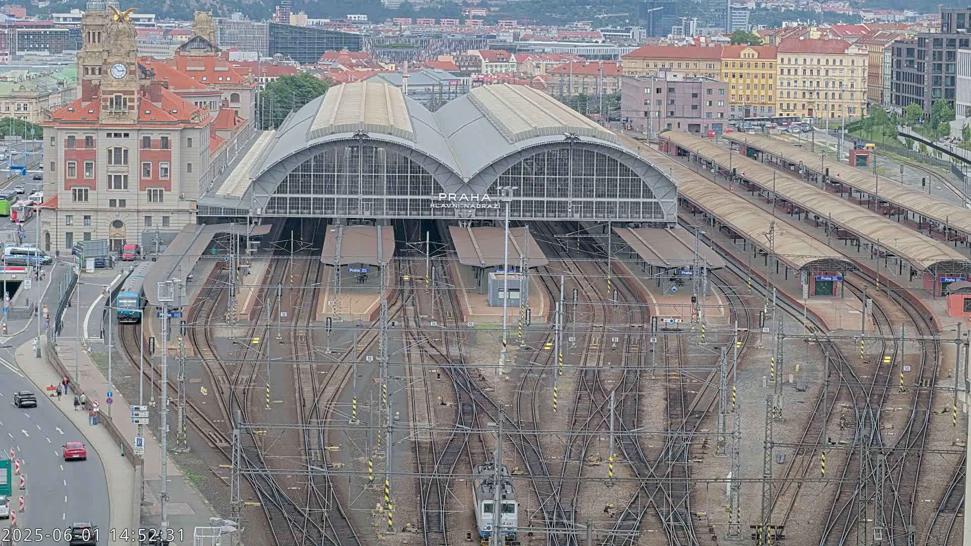 An aerial view shows a large train station with numerous tracks and platforms, surrounded by city buildings under an overcast sky.
