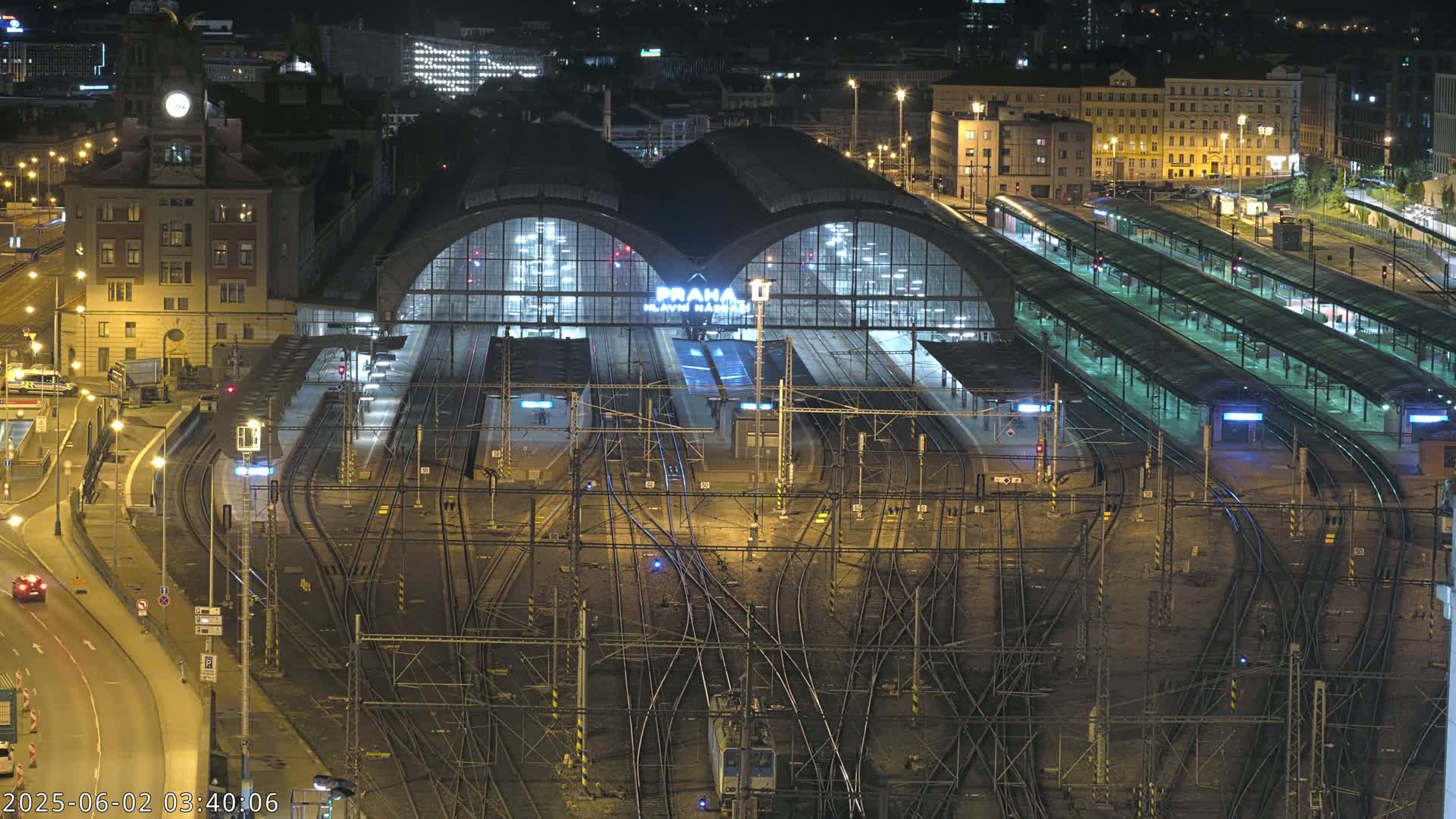 A brightly lit train station at night, with numerous tracks and platforms extending from a large central building, surrounded by city buildings.