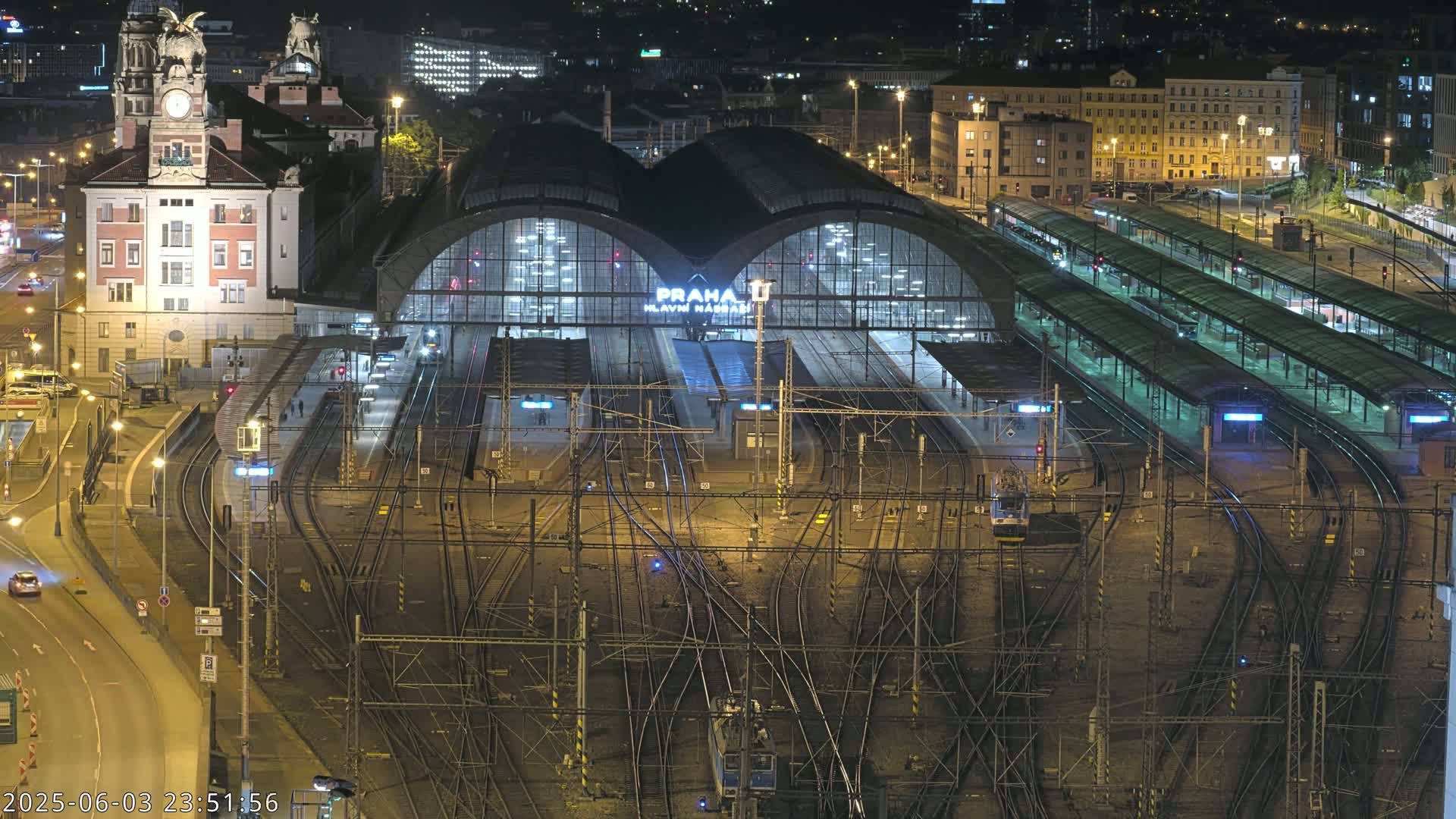 A nighttime aerial view of a large, illuminated train station with numerous tracks and platforms, surrounded by city buildings.