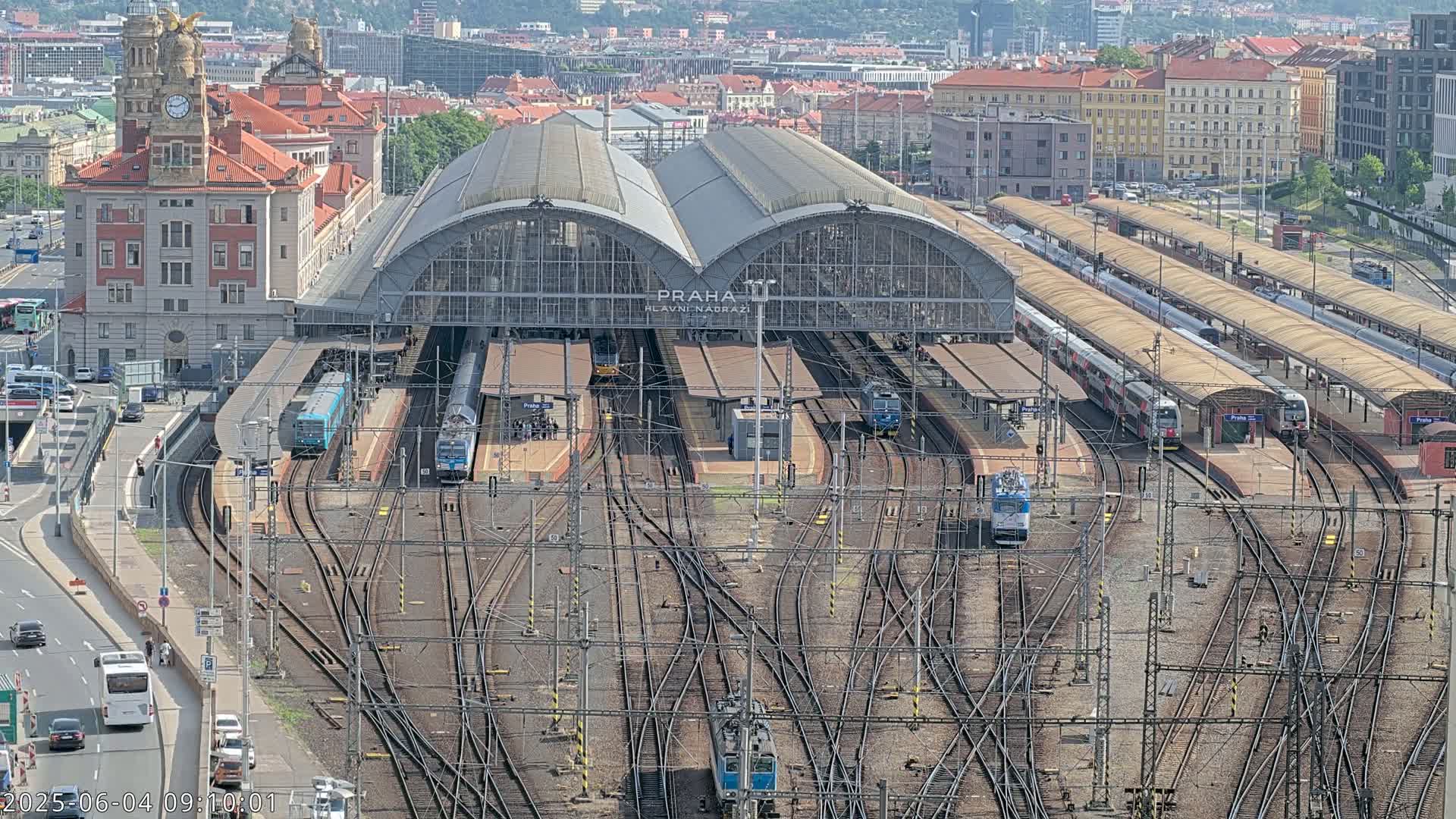 A large train station with numerous trains and tracks, surrounded by city buildings on a sunny day.