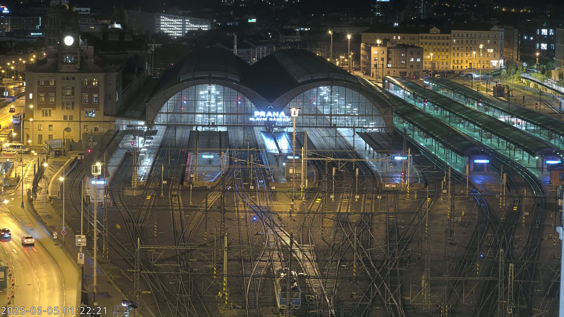 A large, illuminated train station at night, surrounded by numerous train tracks and adjacent buildings.