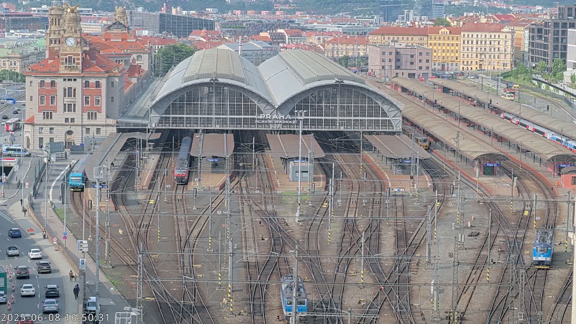 A large train station with numerous tracks and trains, surrounded by city buildings on a sunny day.