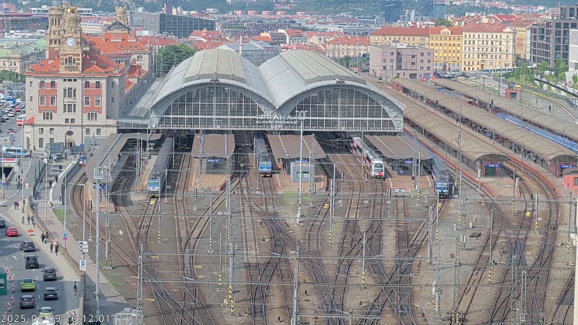 A large train station with numerous tracks and trains, surrounded by city buildings under a clear sky.