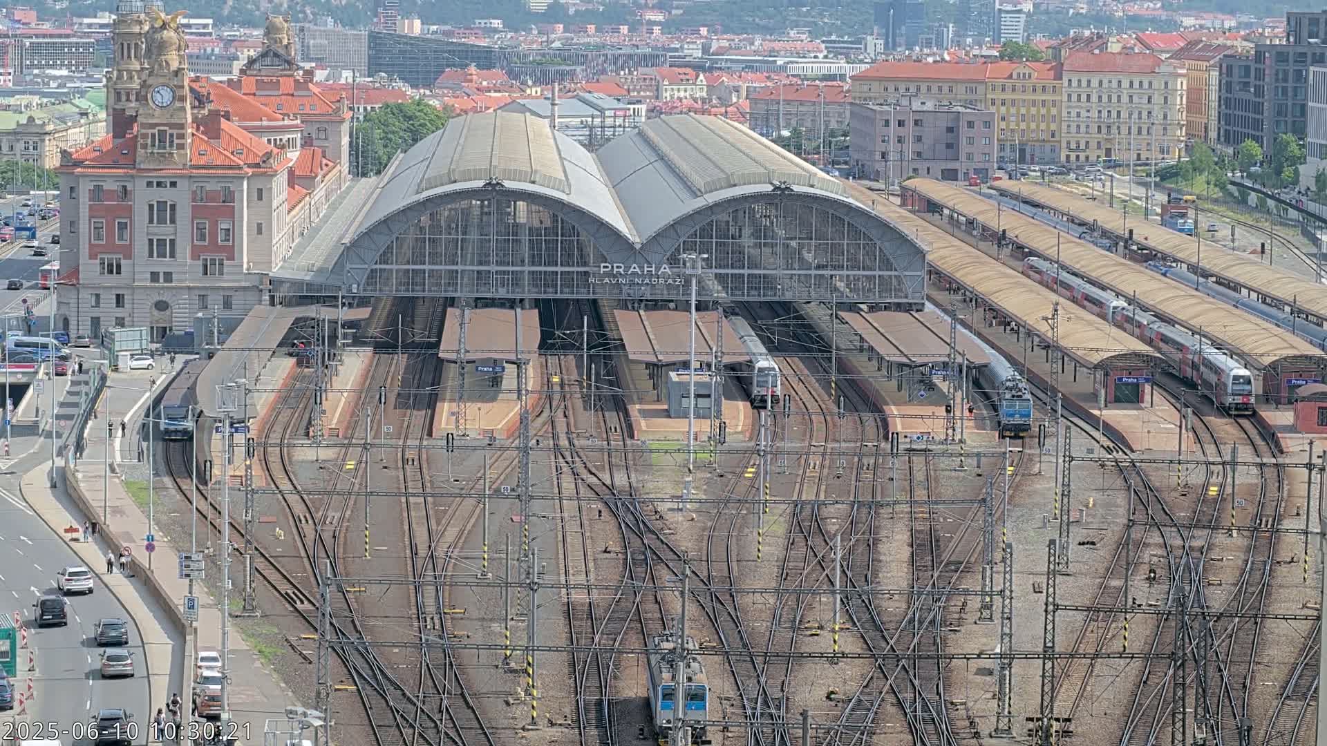 A large train station with numerous tracks and trains, surrounded by city buildings on a sunny day.