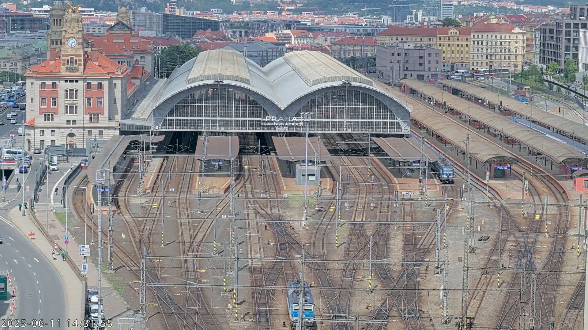 A large train station with numerous tracks and platforms, a main building with arched roofs, and surrounding city buildings is visible on a sunny day.