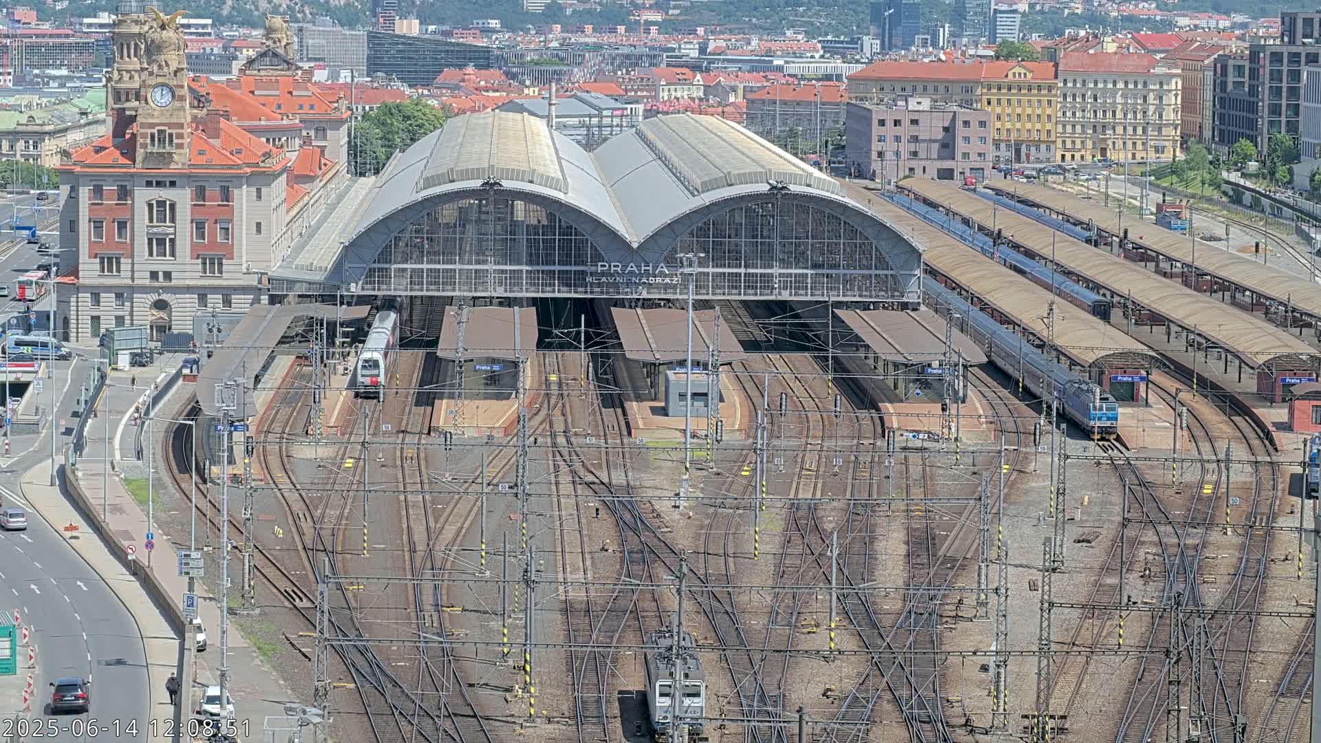 An aerial view shows a large train station with numerous tracks and several trains, under a clear sunny sky.