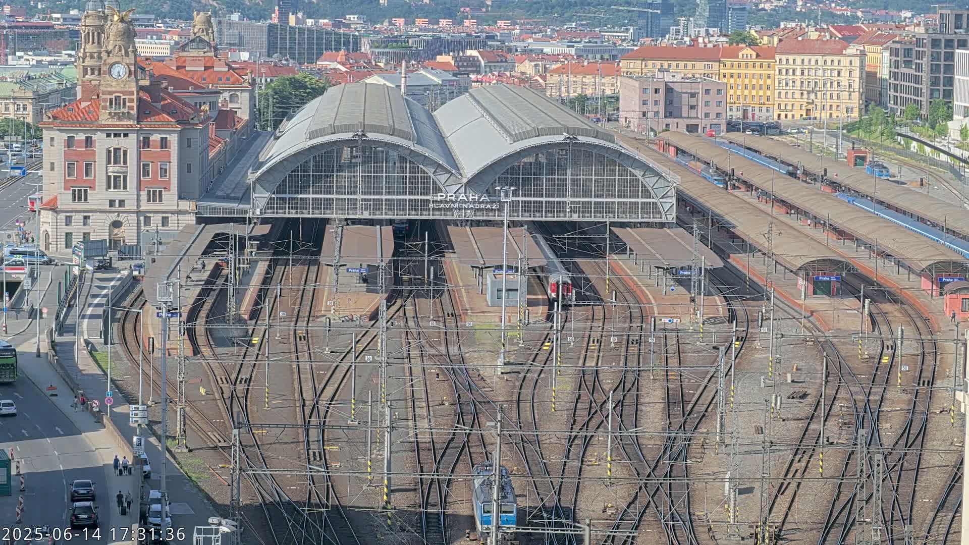 A large train station with numerous tracks and platforms, surrounded by city buildings under a sunny sky.