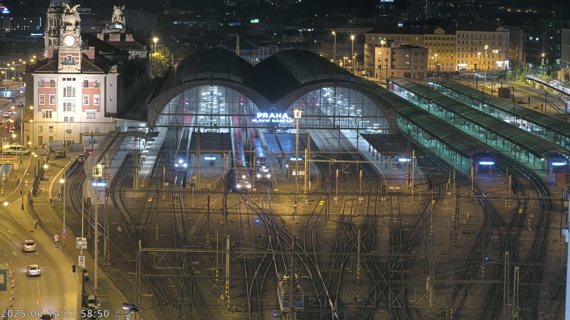 A large train station with numerous tracks and platforms, surrounded by city buildings under a sunny sky.