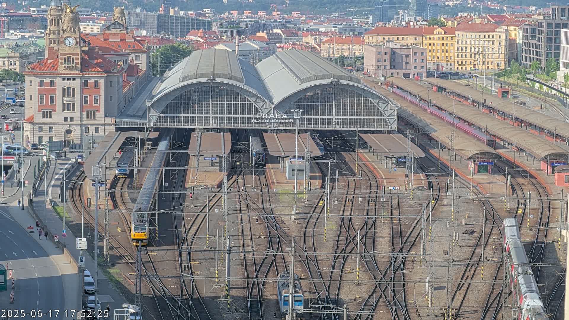 A large train station with numerous tracks and trains, under a clear sky, is viewed from above.