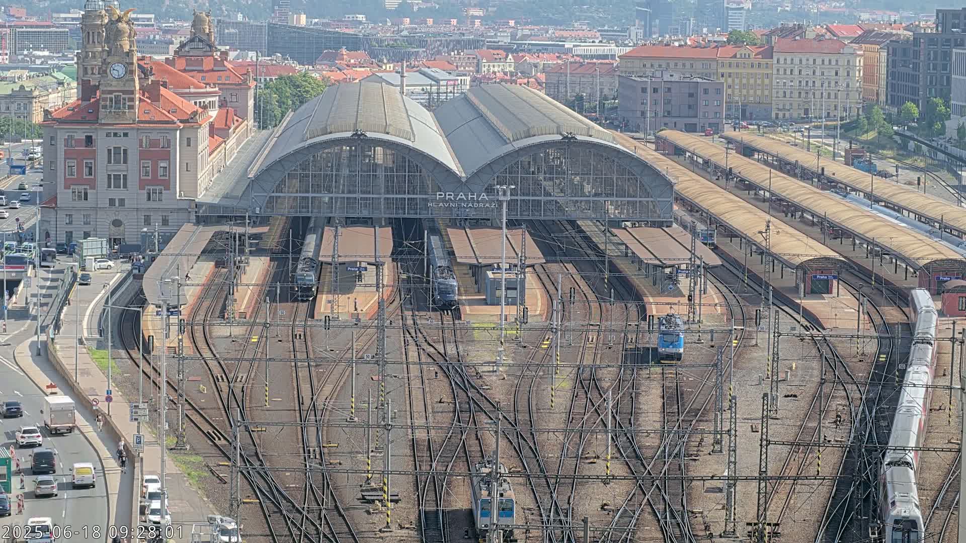 A large train station with numerous tracks and trains, a large building, and surrounding city buildings under a sunny sky.