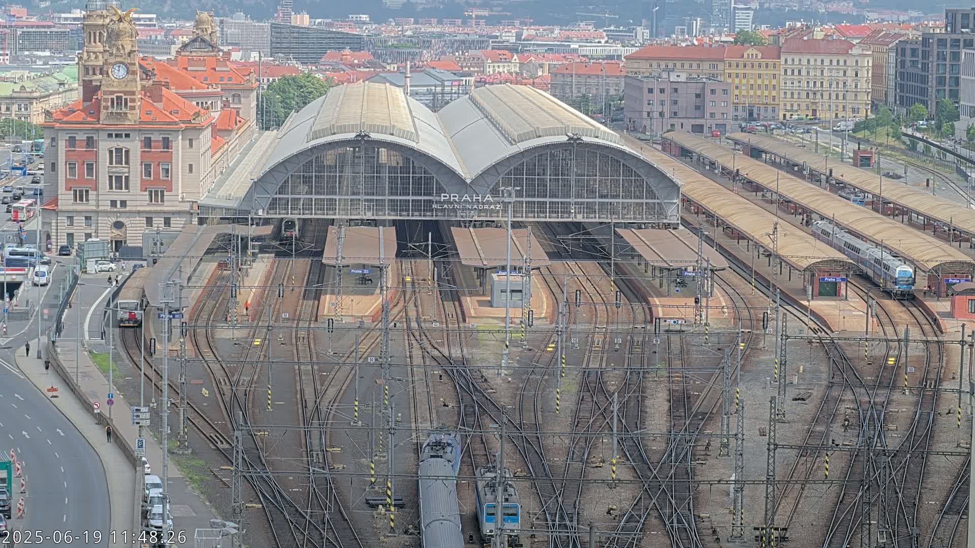 A large train station with numerous tracks and platforms, a large brick building, and several trains visible under a mostly clear sky.