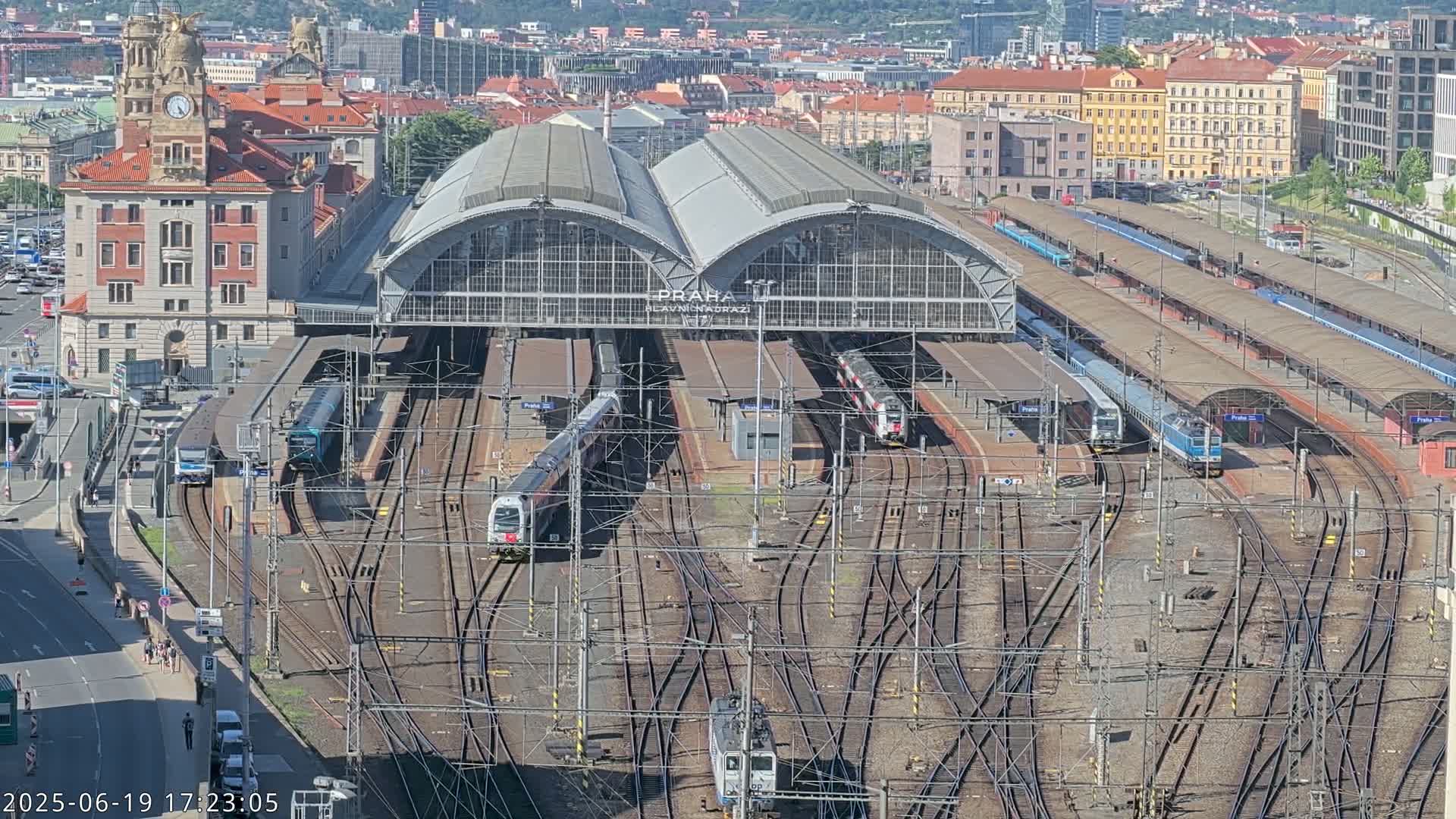 A large train station with many tracks and several trains, under a sunny sky, is viewed from above.