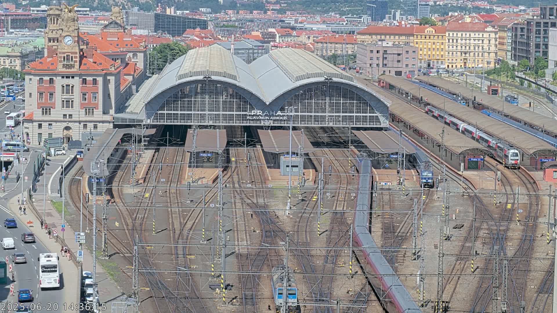 A large train station with numerous tracks and trains, surrounded by city buildings on a sunny day.