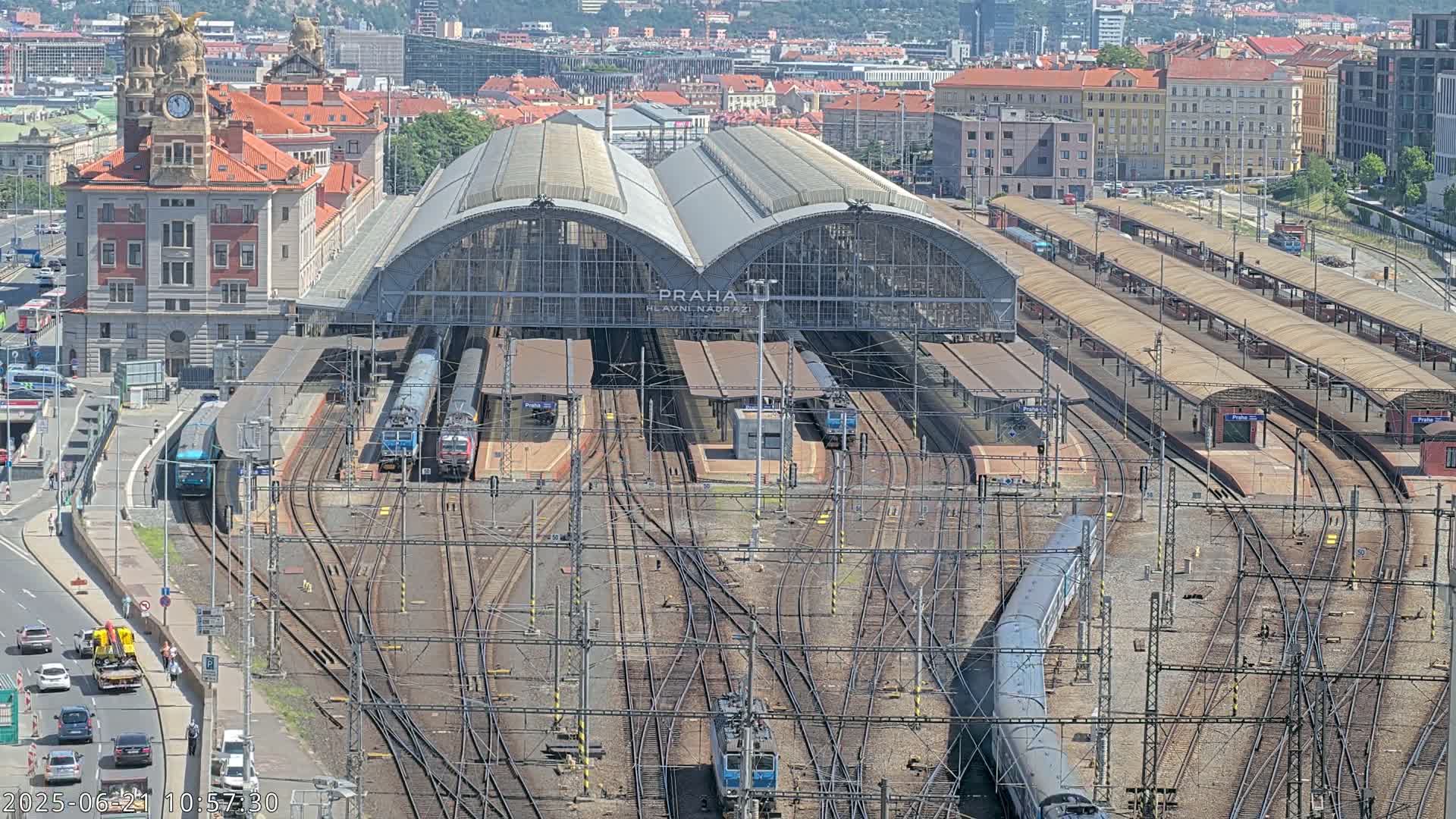 A large train station with numerous tracks and trains, surrounded by city buildings on a sunny day.