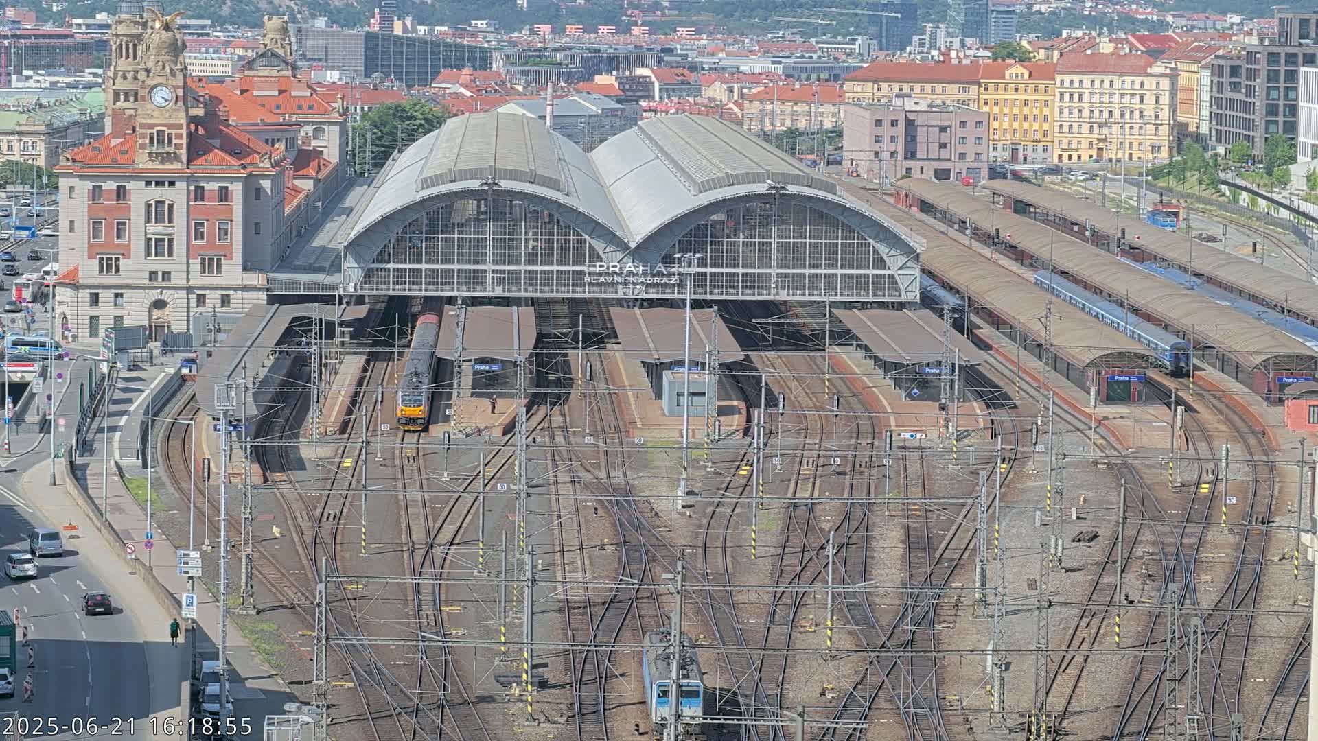 A large train station with numerous tracks and trains, surrounded by city buildings on a sunny day.