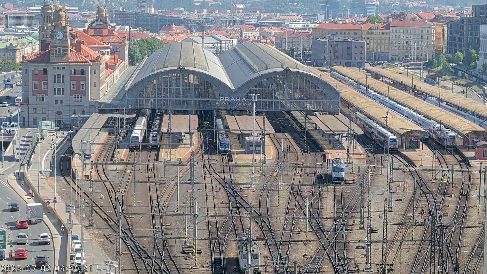 A large train station with many tracks, trains, and platforms is viewed from above on a sunny day.