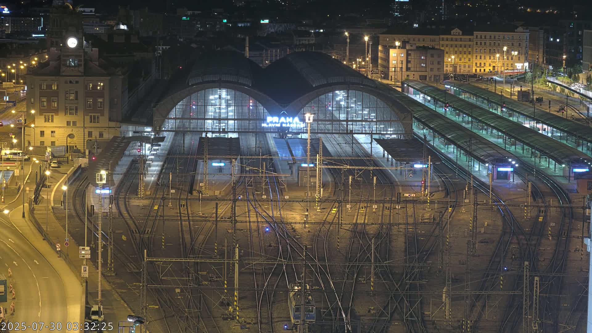 A large train station with many tracks, trains, and platforms is viewed from above on a sunny day.