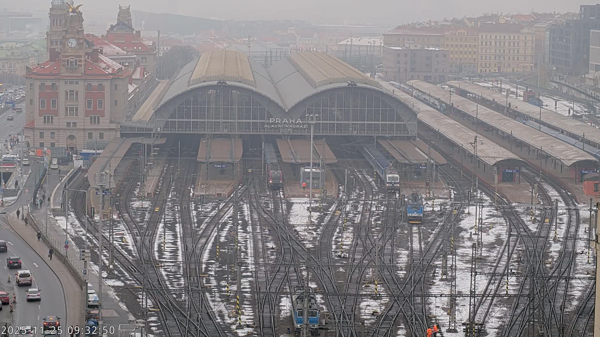 A wide aerial shot on an overcast and cold day reveals a vast train station with numerous tracks partially covered in snow and slush, several trains at platforms, and a grand clock tower building adjacent to a busy road with vehicles and pedestrians.