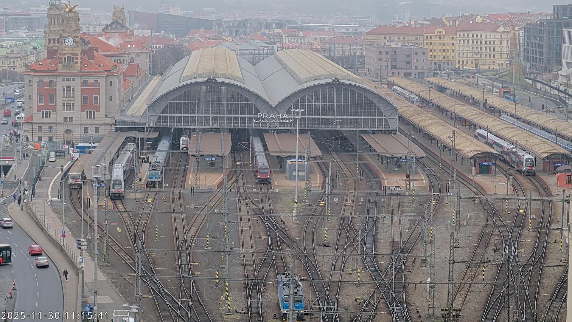 An aerial view reveals a vast train station with numerous tracks and multiple trains, flanked by a grand clock tower building and other city structures, all under an overcast and hazy sky.