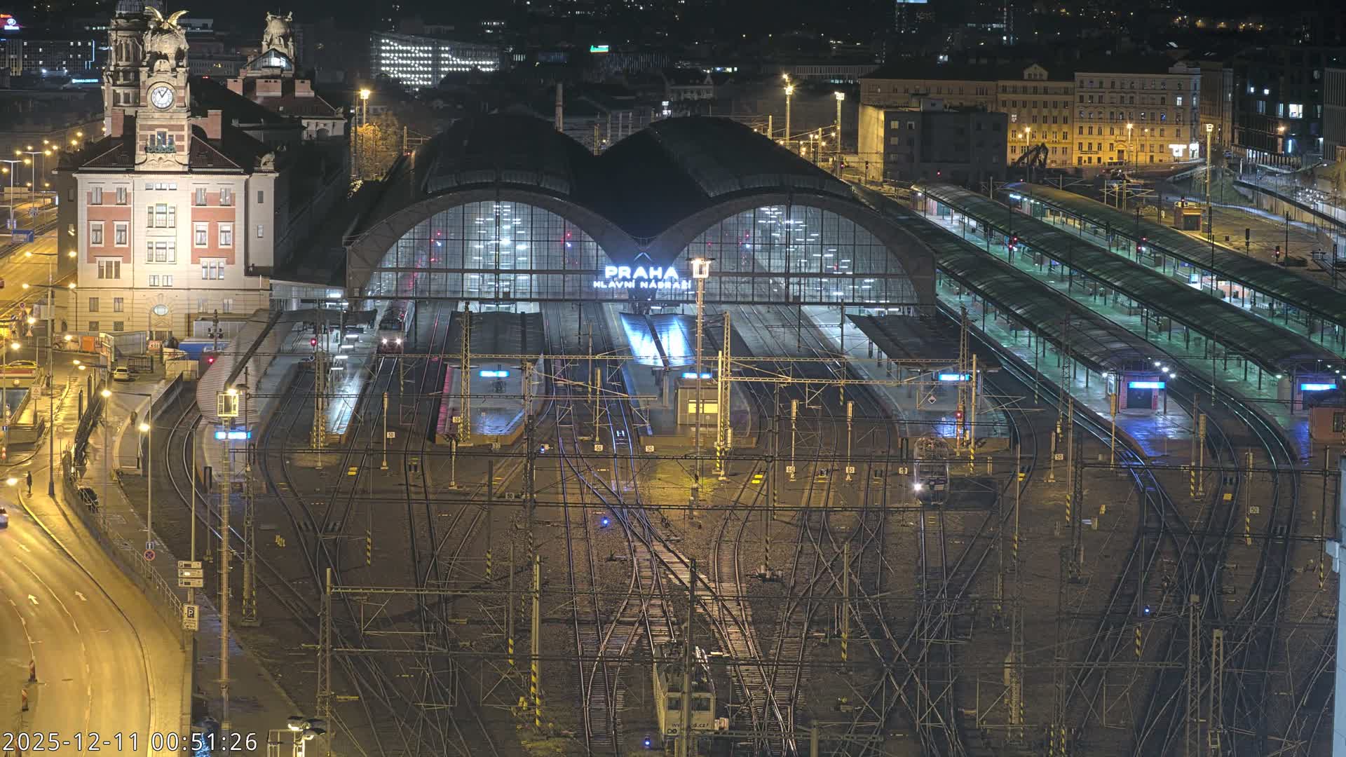 An elevated nighttime view reveals a sprawling, brightly lit train station with numerous tracks and platforms under a large arched roof, surrounded by illuminated city buildings and wet roads, indicative of rainy conditions.