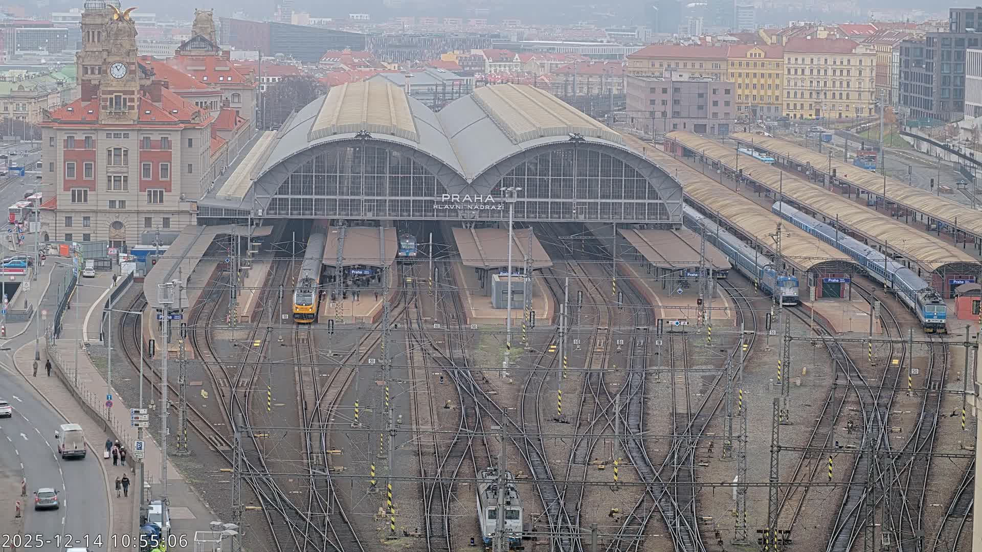 An elevated nighttime view reveals a sprawling, brightly lit train station with numerous tracks and platforms under a large arched roof, surrounded by illuminated city buildings and wet roads, indicative of rainy conditions.