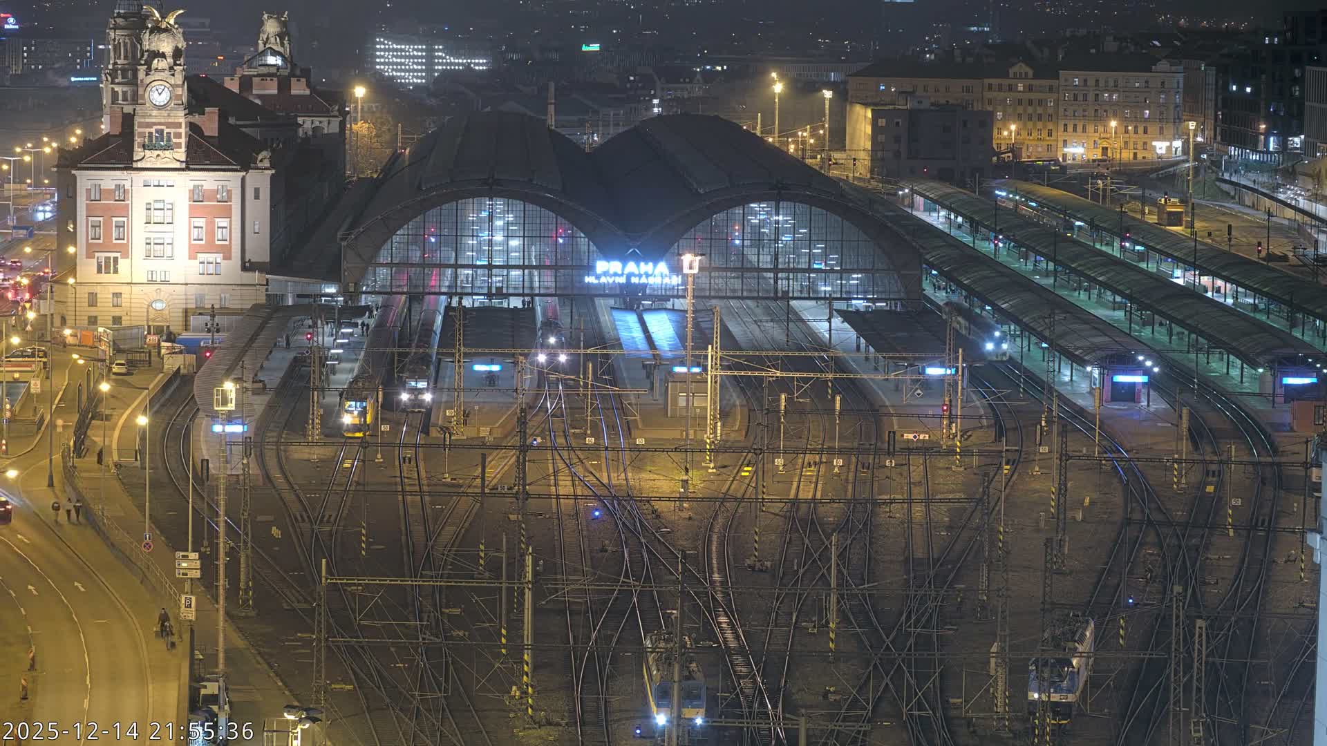 An elevated nighttime view reveals a sprawling, brightly lit train station with numerous tracks and platforms under a large arched roof, surrounded by illuminated city buildings and wet roads, indicative of rainy conditions.
