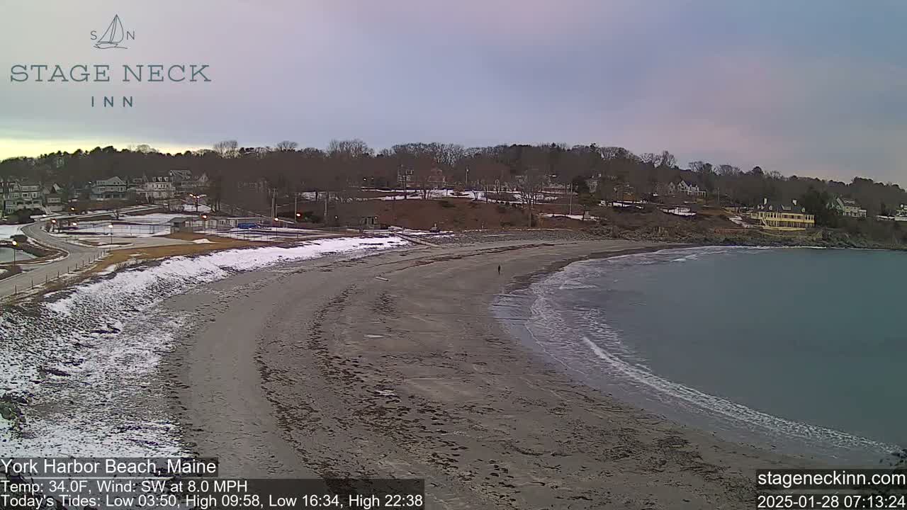 A mostly sandy beach with some snow, gentle waves lapping the shore, and a few buildings in the background under an overcast sky.