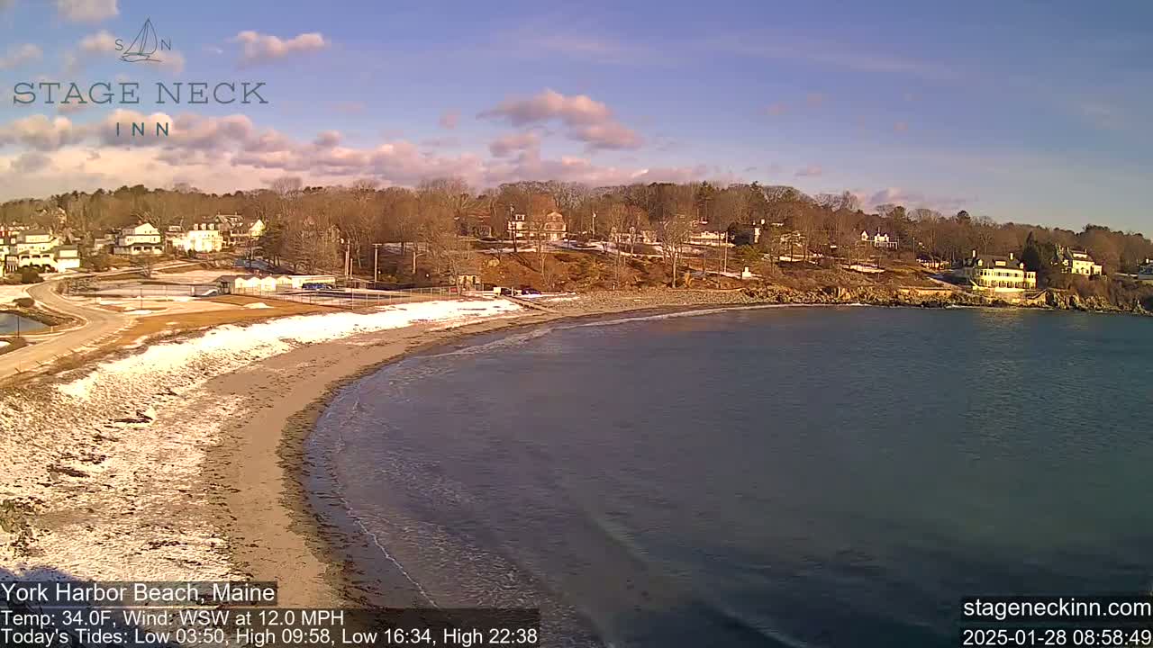 A sandy beach with patches of snow curves into a calm, dark-blue bay under a mostly clear, sunny sky, and houses sit on a hillside overlooking the beach.