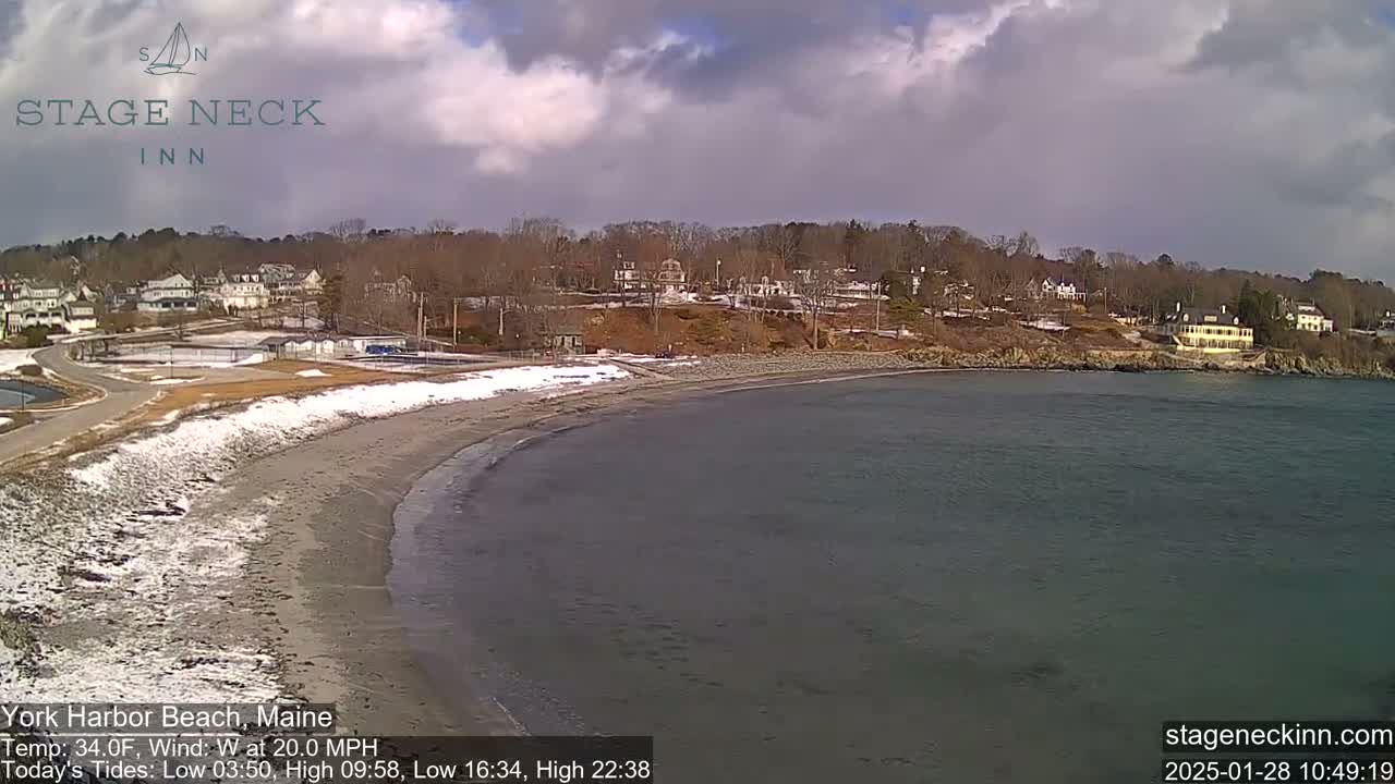 A partly cloudy day shows a snow-dusted beach curving into a calm bay, with buildings and trees on a hillside beyond.