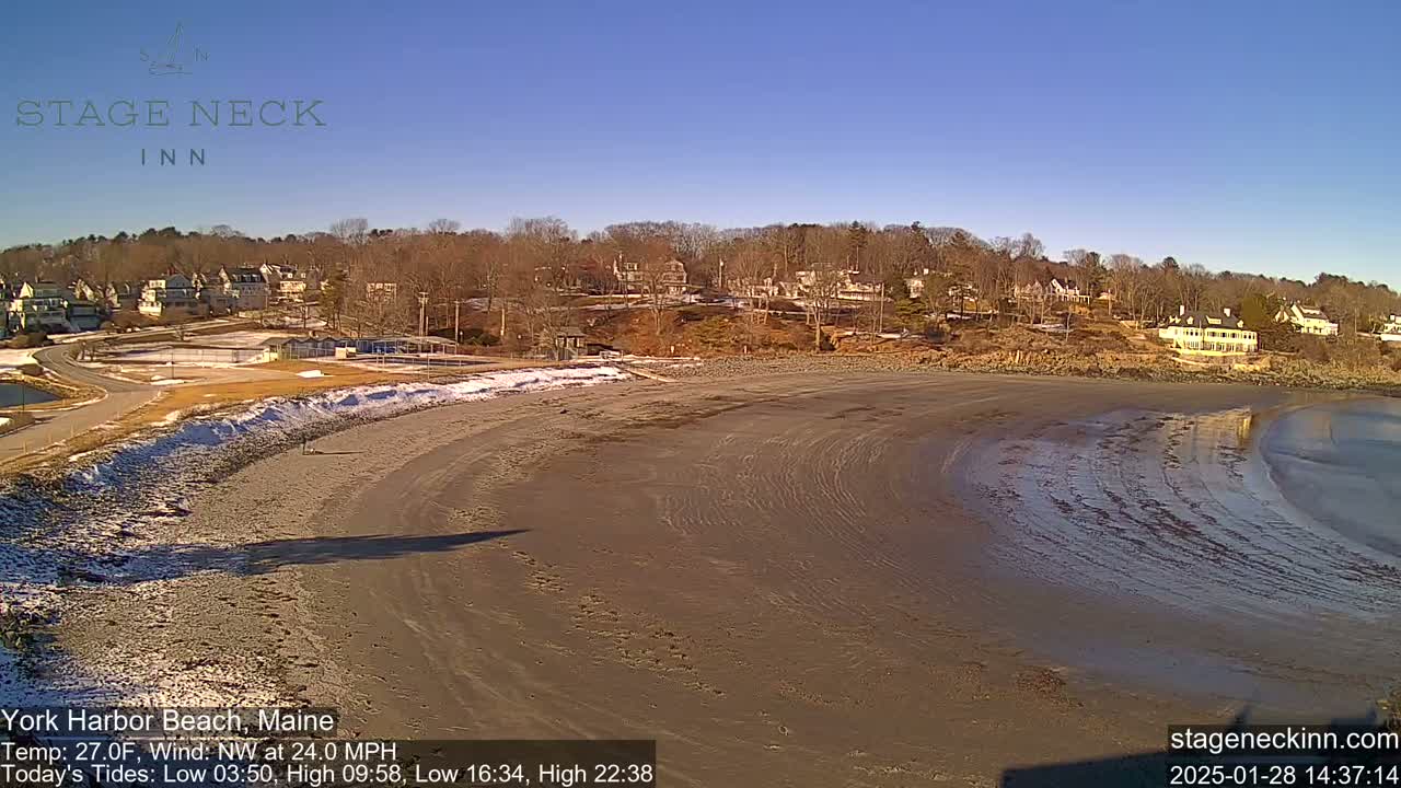 A sandy beach curves into a calm body of water under a clear blue sky, with a residential area visible in the background.