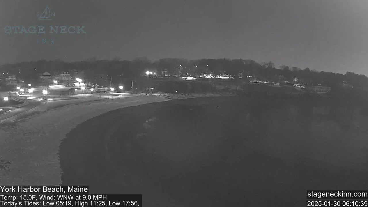 A nighttime view of a dark beach and a snow-covered parking area next to a line of buildings, under a dark sky with a 15°F temperature and a 9 mph wind from the west-northwest.