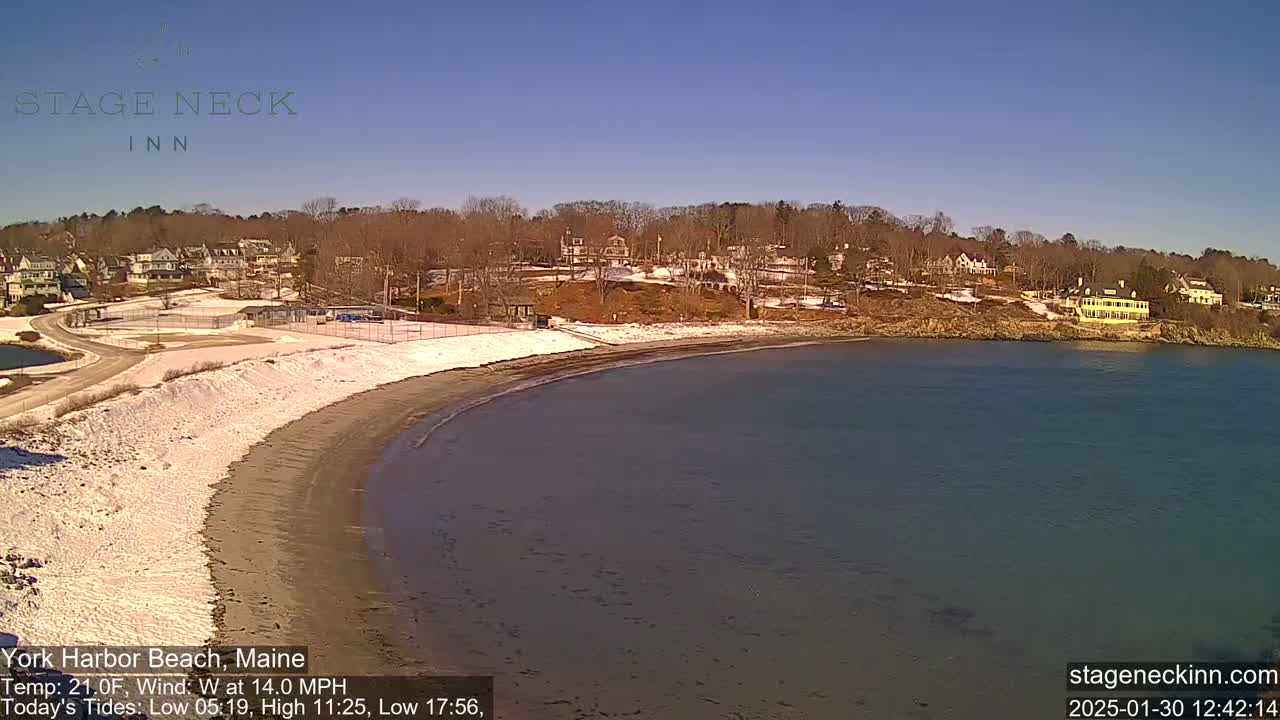 A partially snow-covered beach curves into a calm body of water under a clear blue sky, with houses and trees visible on the far shore.