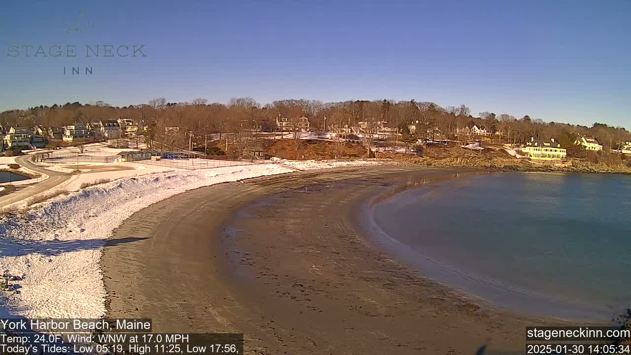 A sandy beach curves into a calm bay, with a snow-dusted shoreline and buildings visible in the background under a clear blue sky.