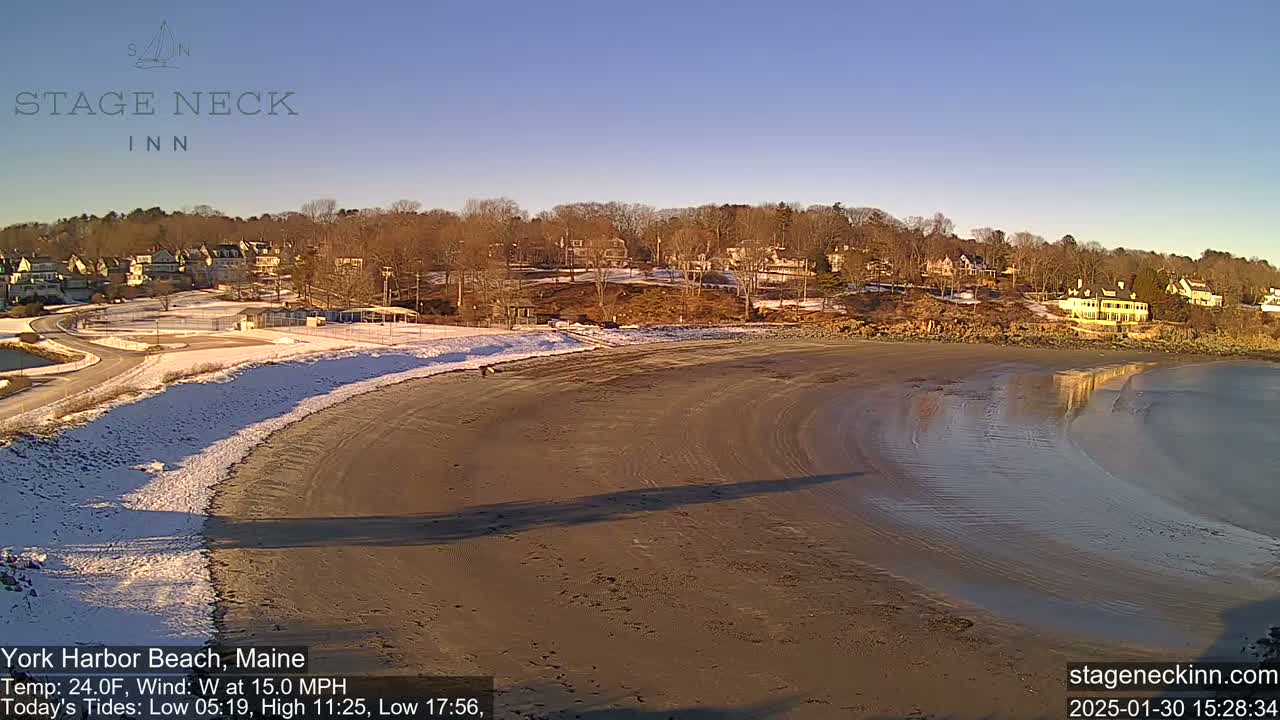 A sandy beach with some snow partially covers the shore next to a calm, shallow body of water, with houses and trees visible in the background under a clear, sunny sky.