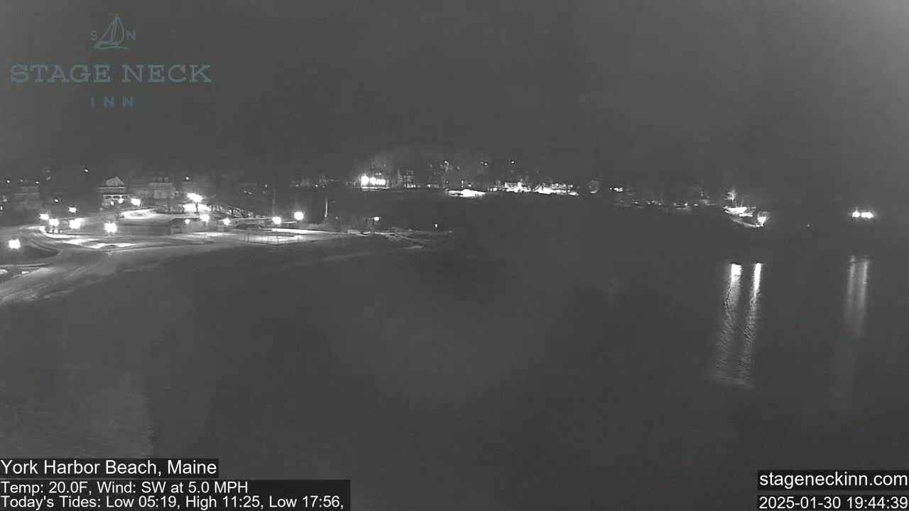A nighttime view of a snow-covered beach and dark water, with faintly visible lights from buildings in the distance under a dark sky.