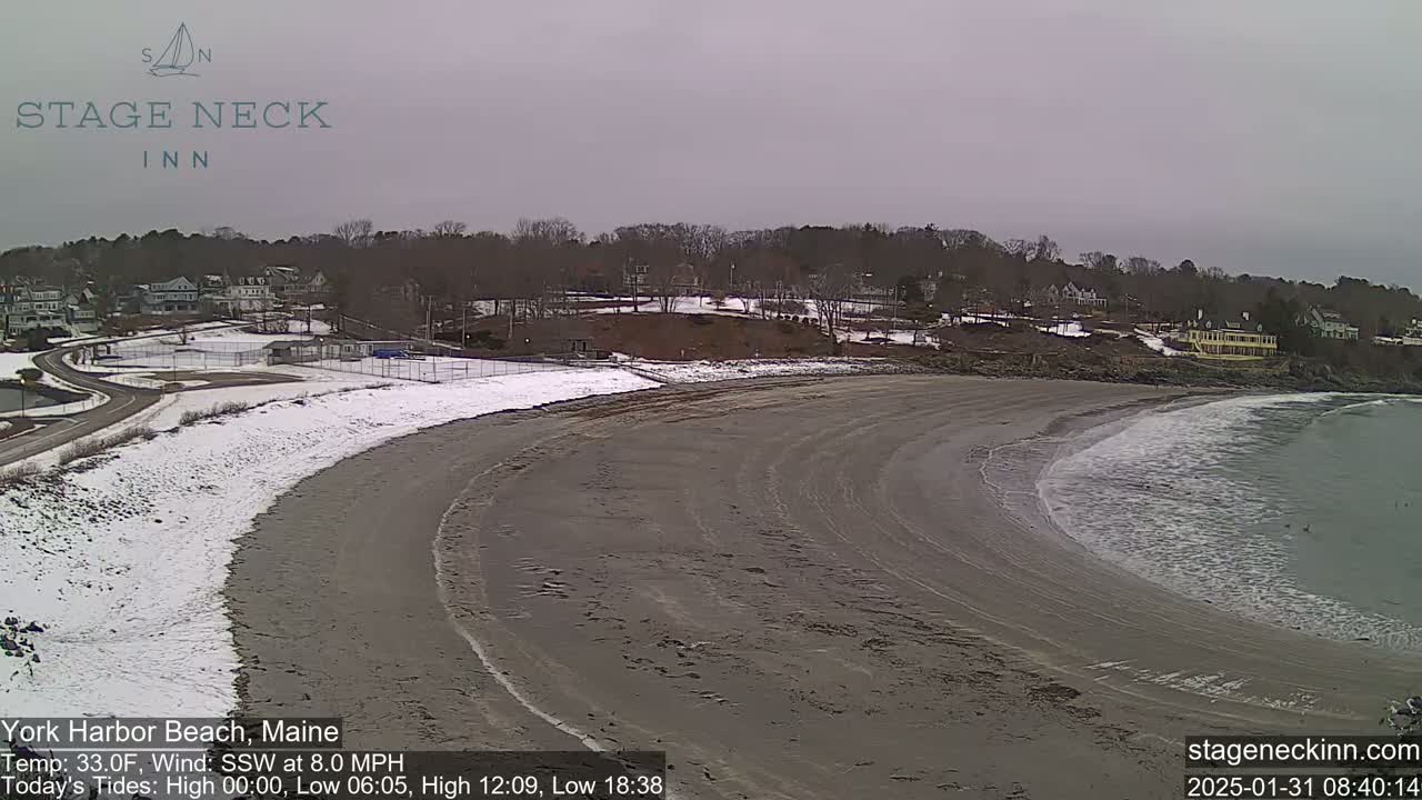 A sandy beach with some snow, gently sloping into a calm ocean under an overcast sky.