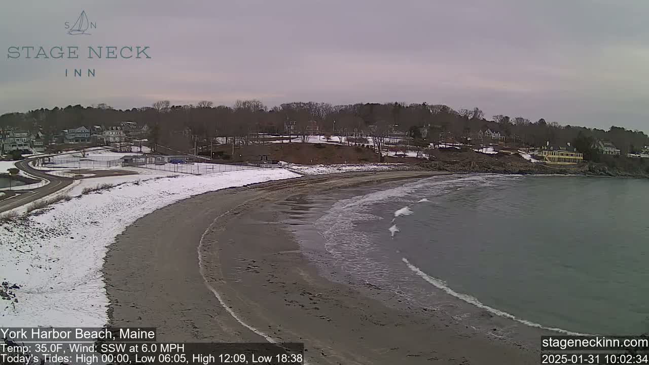 A partially snow-covered sandy beach curves gently into a calm, gray ocean under an overcast sky.