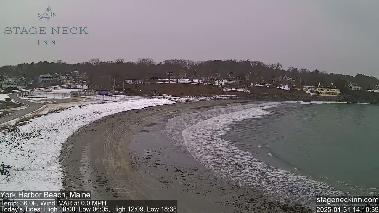 A sandy beach partially covered in snow slopes down to a calm, gray ocean with small waves under an overcast sky.