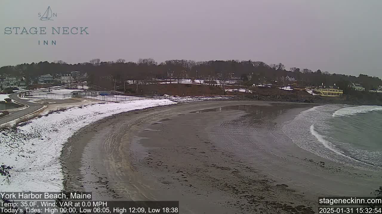 A partly snowy beach with calm waves and a residential area in the background under an overcast sky.