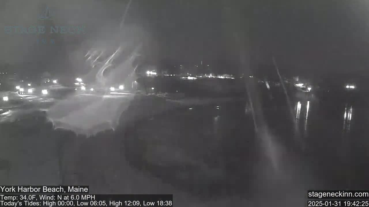 A nighttime, snowy scene shows a road with cars driving through a lightly snow-covered area near a body of water.