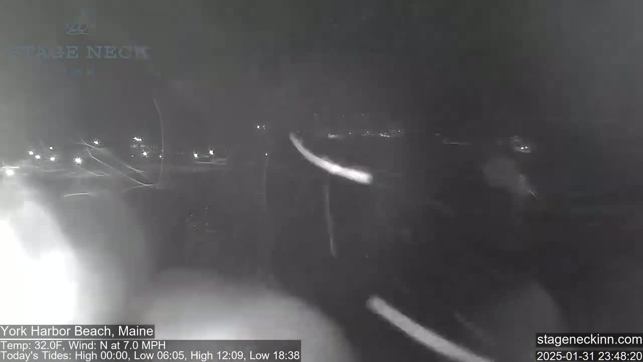 A nighttime, snowy scene shows blurred headlights of vehicles on a road, with a light wind.