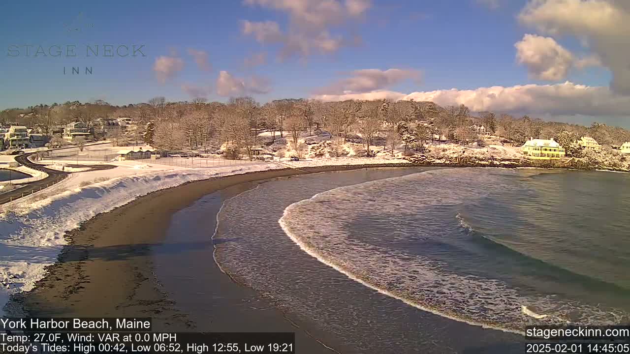 A snow-covered beach with a calm ocean and a few small waves gently lapping the shore, under a mostly sunny sky with some clouds.