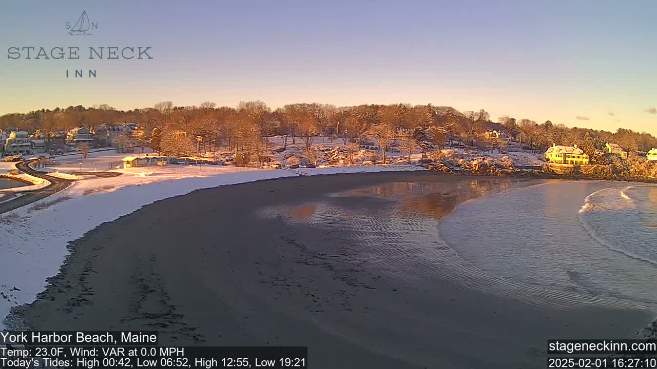 A snow-covered beach with a calm ocean, showing small waves gently lapping the shore, under a clear, bright sky at sunrise.