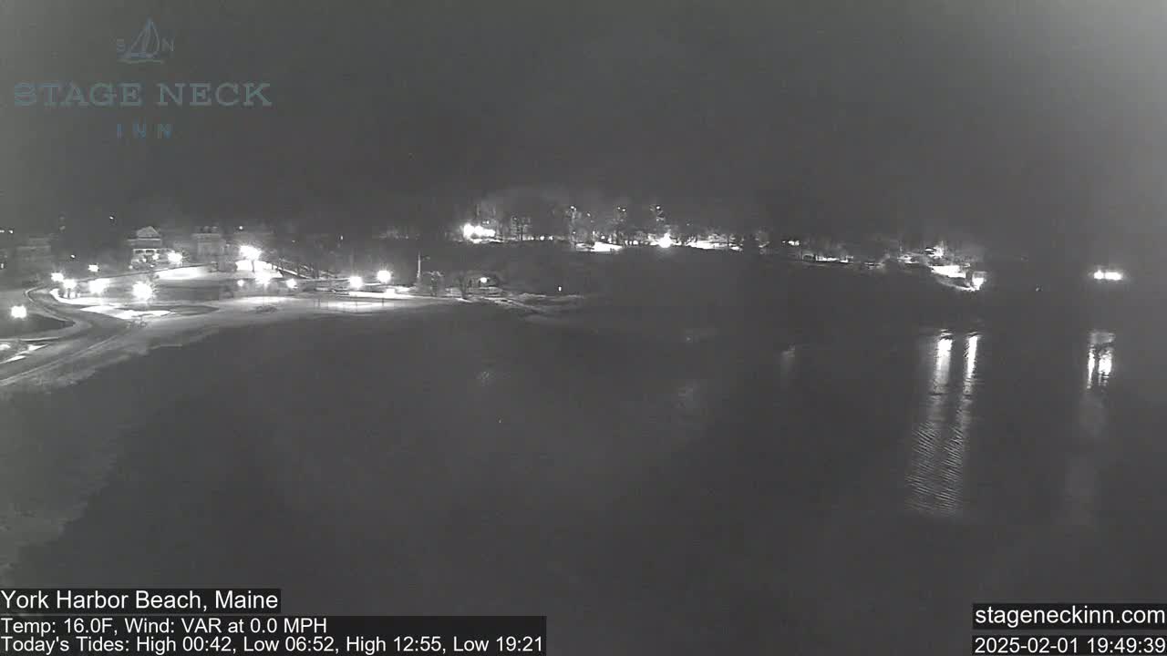 A nighttime, snow-covered beach scene shows a calm body of water reflecting lights from buildings on the shore.