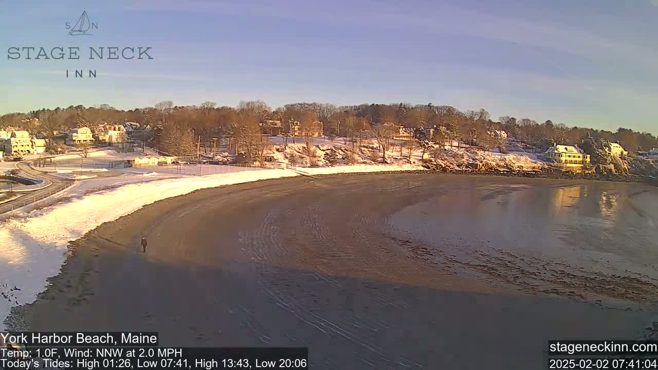 A person walks along a sandy beach partially covered in snow, with a calm body of water reflecting the sunlight, and houses covered in snow on a hillside in the background under a clear sky.