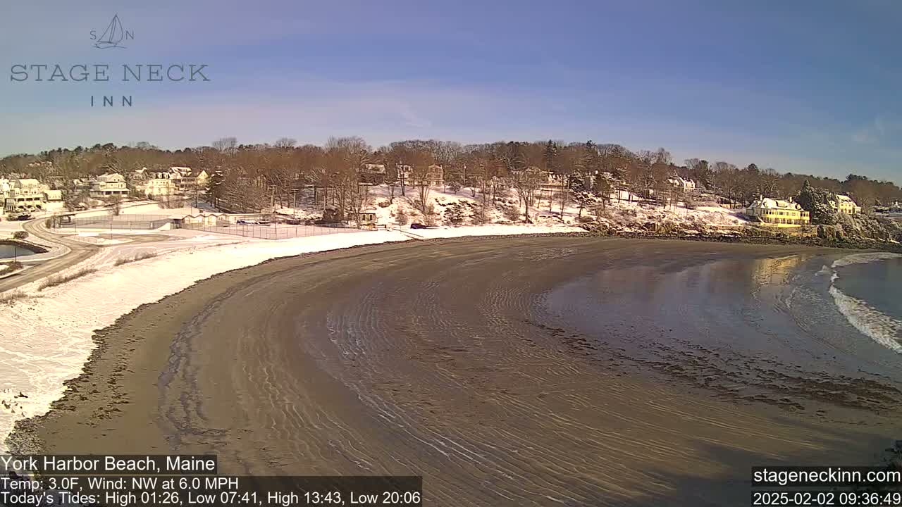 A snow-covered beach curves along a coastline, with a calm sea and houses visible on a hillside in the background under a clear sky.