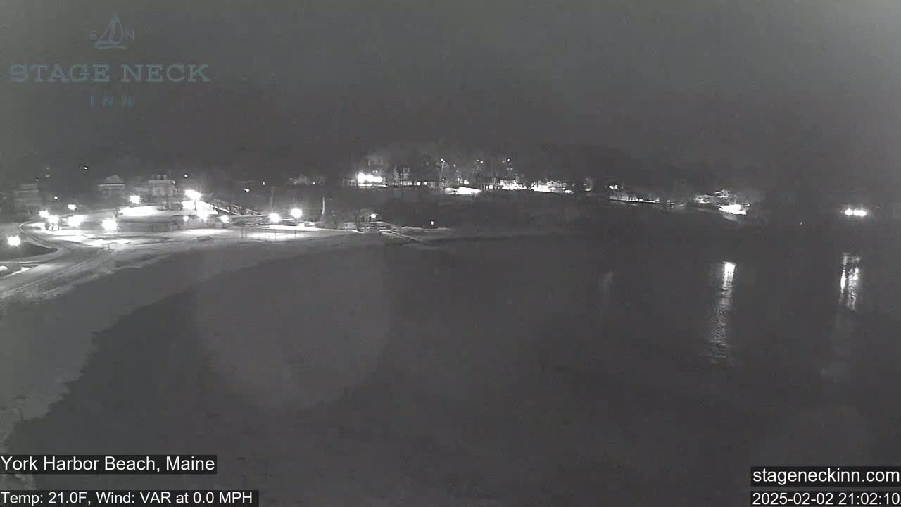 A nighttime, snow-covered beach scene with calm water and faintly lit buildings in the background under a dark, possibly cloudy sky.