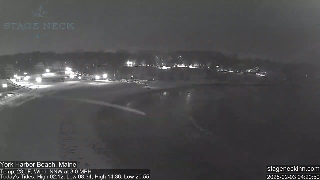 A snow-covered beach at night is dimly lit by streetlights, with a dark body of water visible in the foreground under a dark sky.