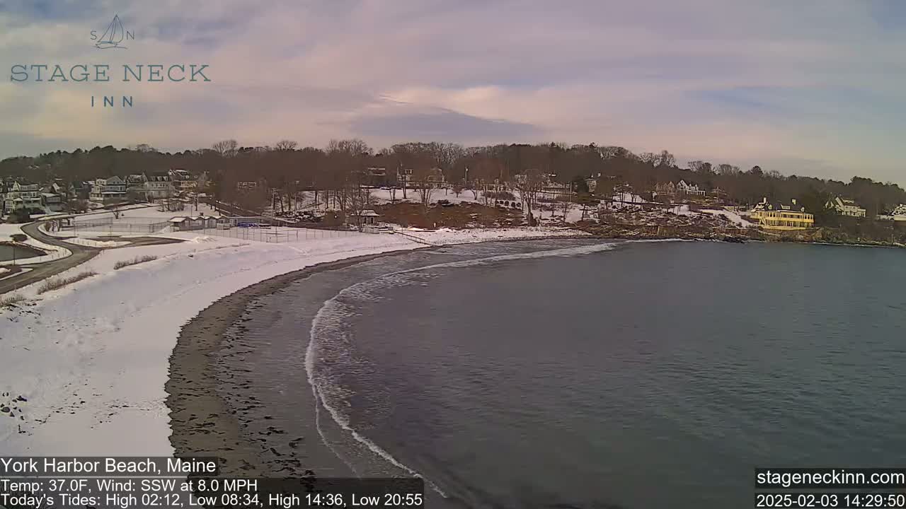 A snow-dusted beach curves into a dark gray ocean with gentle waves under a cloudy sky.